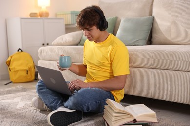 Photo of Teenage student with cup of drink using laptop while studying on floor at home