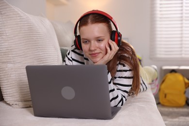 Photo of Teenage student in headphones using laptop while studying on sofa at home
