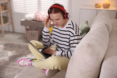 Photo of Teenage student in headphones using laptop while studying on sofa at home