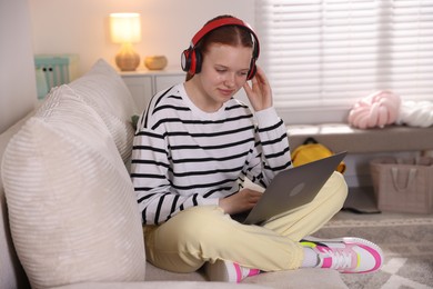Photo of Teenage student in headphones using laptop while studying on sofa at home