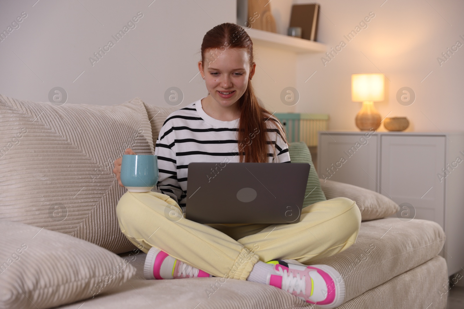 Teenage student with cup of drink using laptop while studying on sofa at home Photo of Teenage student with cup of drink using laptop while studying on sofa at home