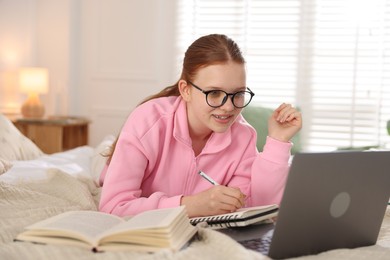 Photo of Teenage student taking notes while studying near laptop on bed at home