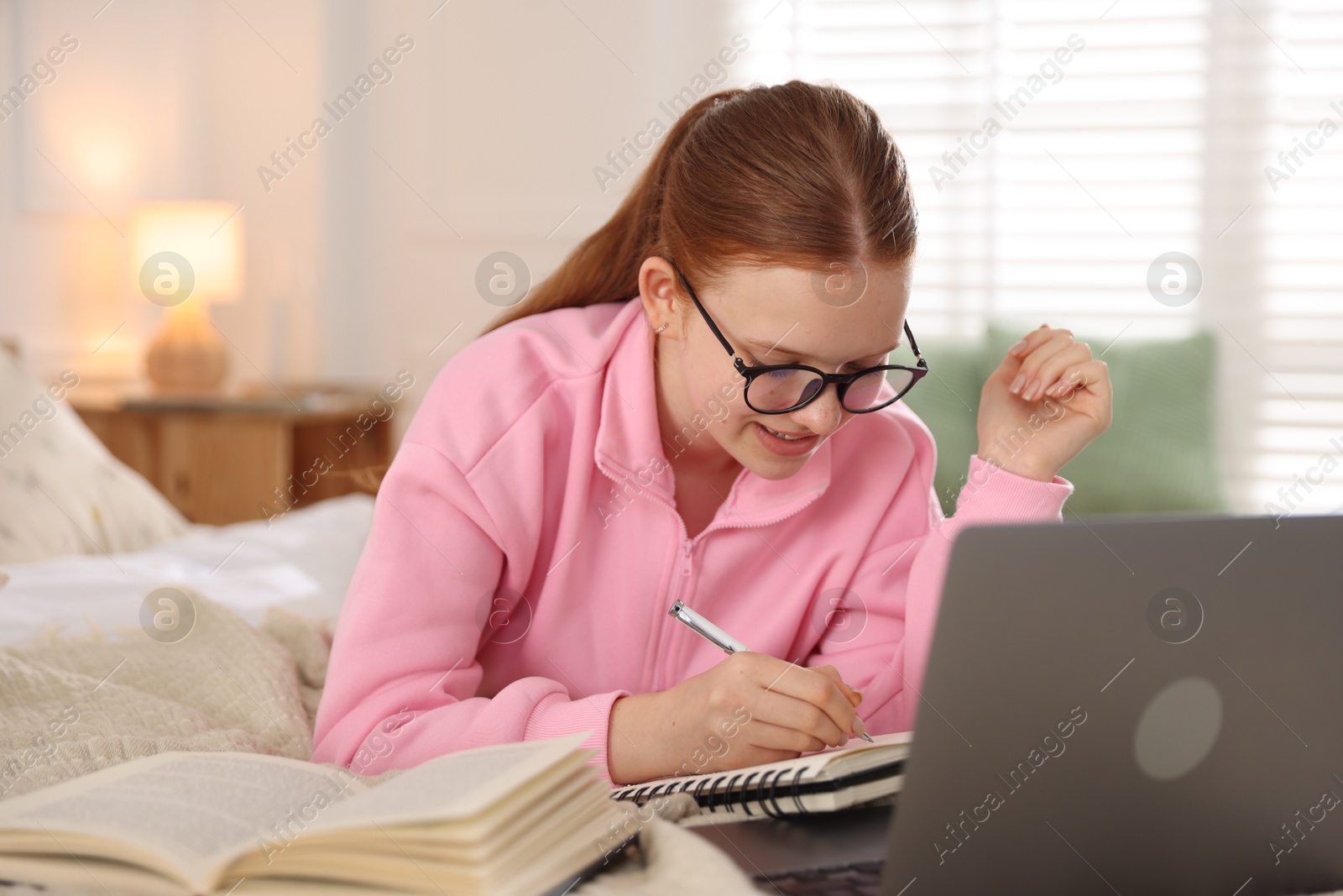 Teenage student taking notes while studying near laptop on bed at home Photo of Teenage student taking notes while studying near laptop on bed at home