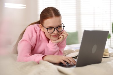 Photo of Teenage student in glasses using laptop while studying on bed at home