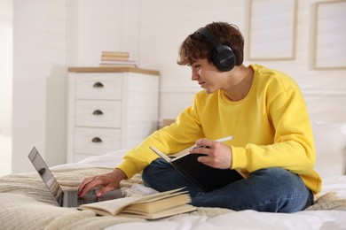 Photo of Teenage student in headphones using laptop while studying on bed at home