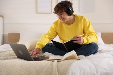 Photo of Teenage student in headphones using laptop while studying on bed at home