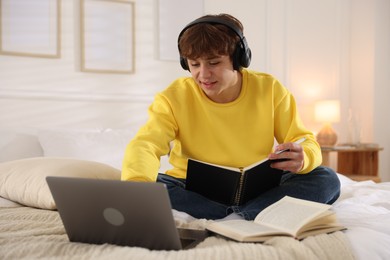 Photo of Teenage student in headphones using laptop while studying on bed at home