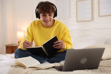 Photo of Teenage student in headphones using laptop while studying on bed at home