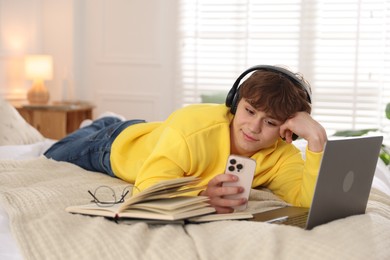 Photo of Teenage student in headphones using smartphone while studying on bed at home
