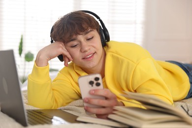 Photo of Teenage student in headphones using smartphone while studying on bed at home