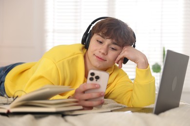 Photo of Teenage student in headphones using smartphone while studying on bed at home