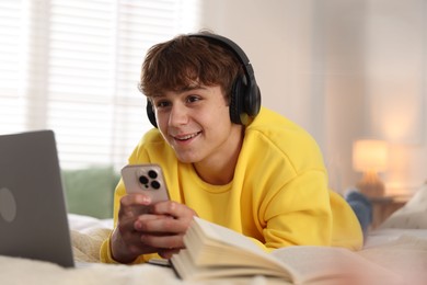 Photo of Teenage student in headphones using smartphone while studying on bed at home
