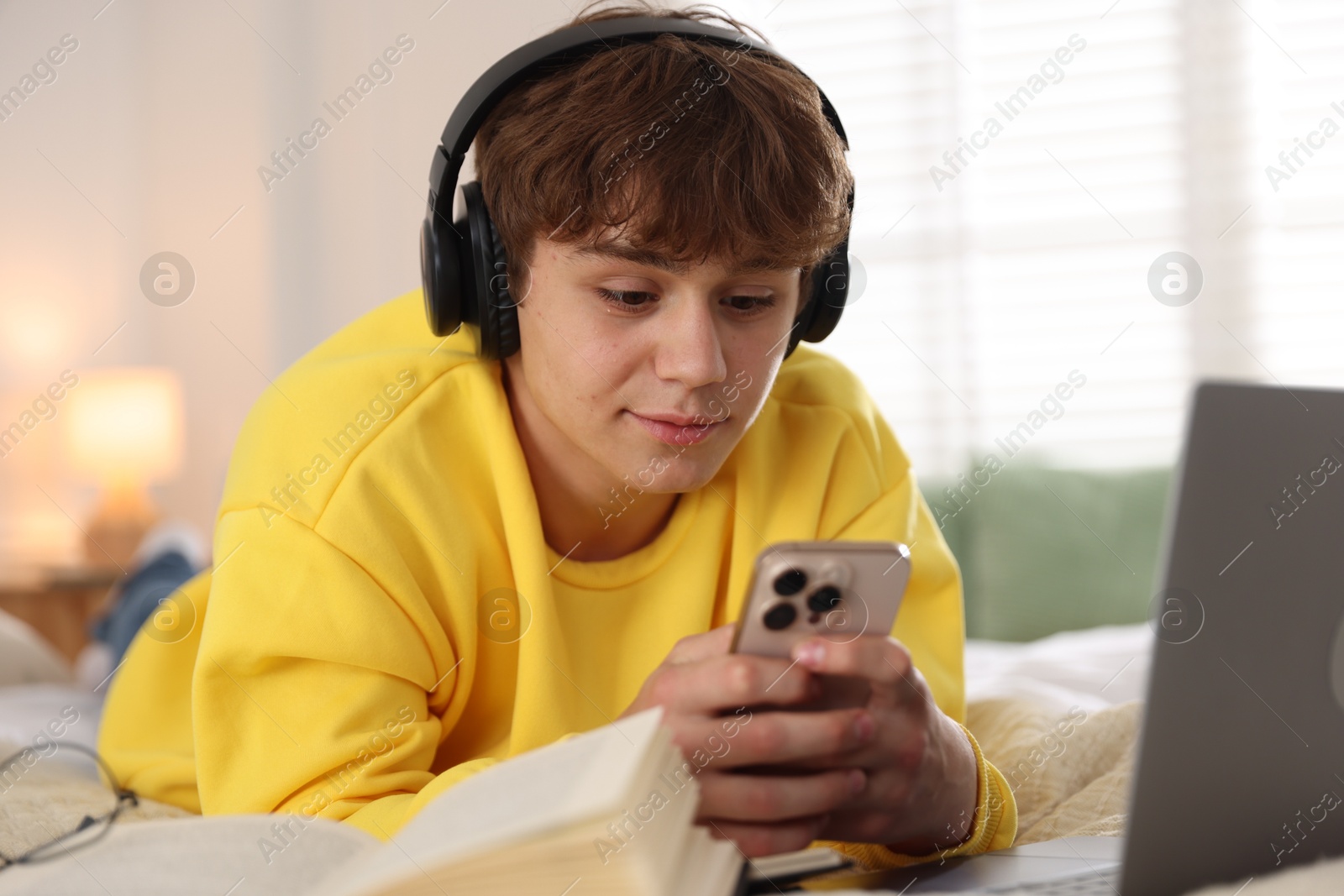 Teenage student in headphones using smartphone while studying on bed at home Photo of Teenage student in headphones using smartphone while studying on bed at home