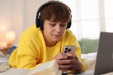 Photo of Teenage student in headphones using smartphone while studying on bed at home