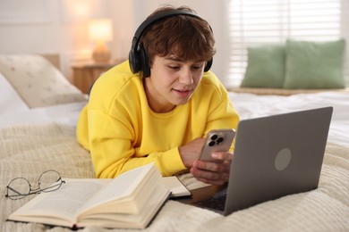 Photo of Teenage student in headphones using smartphone while studying on bed at home
