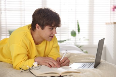 Photo of Teenage student taking notes while studying near laptop on bed at home