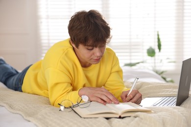 Photo of Teenage student taking notes while studying near laptop on bed at home