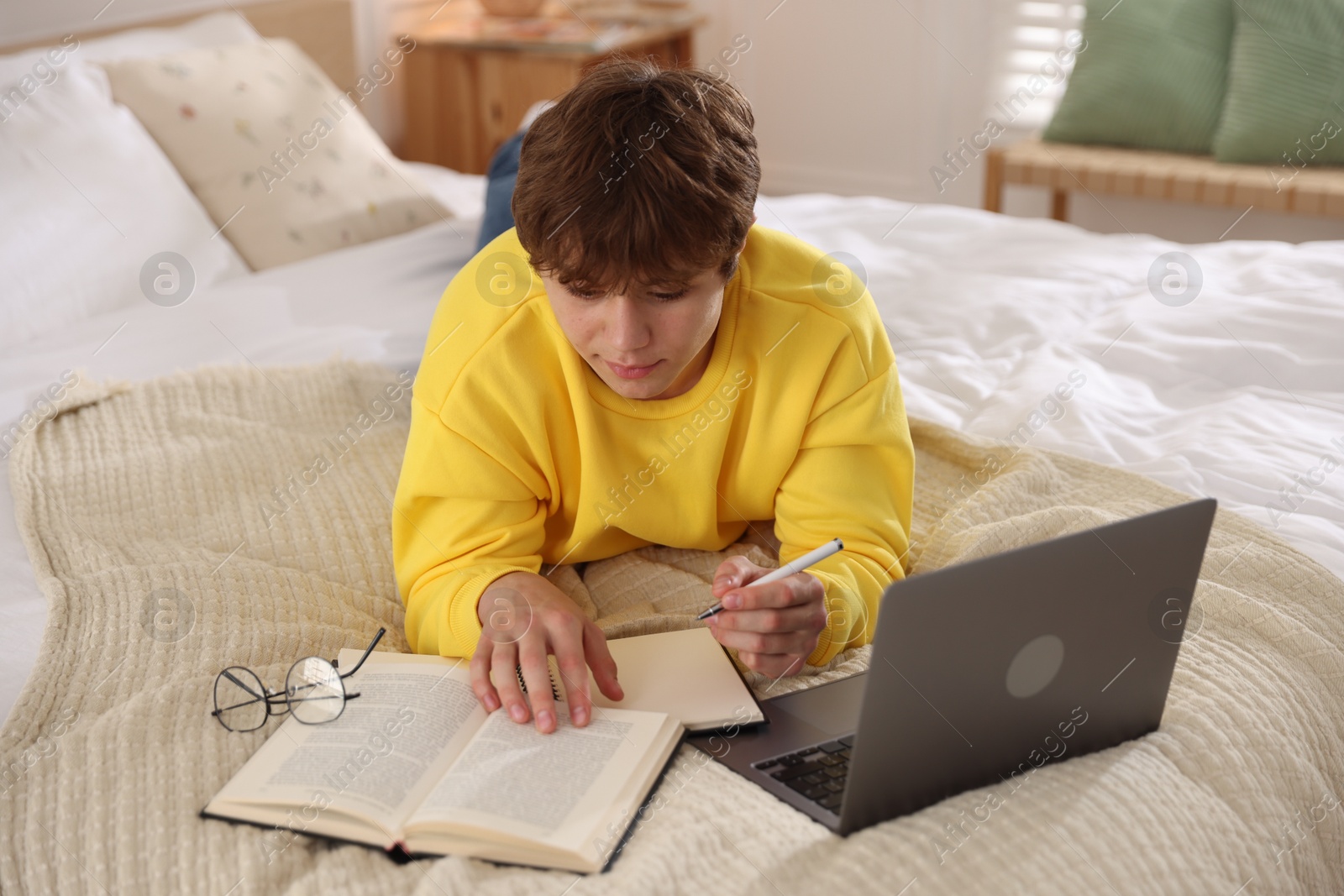 Photo of Teenage student taking notes while studying near laptop on bed at home