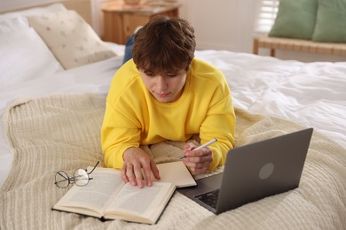 Photo of Teenage student taking notes while studying near laptop on bed at home
