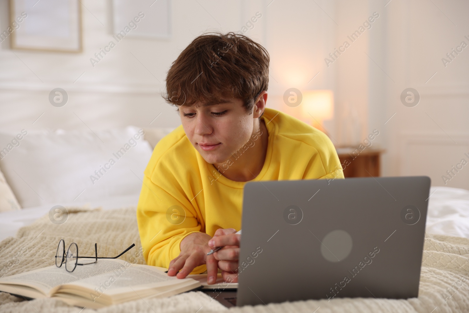 Teenage student taking notes while studying near laptop on bed at home Photo of Teenage student taking notes while studying near laptop on bed at home