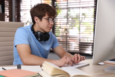 Photo of Teenage student with headphones using computer while studying at table indoors