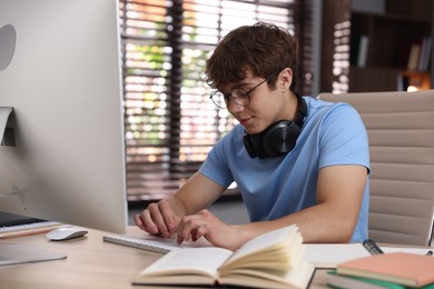 Photo of Teenage student with headphones using computer while studying at table indoors