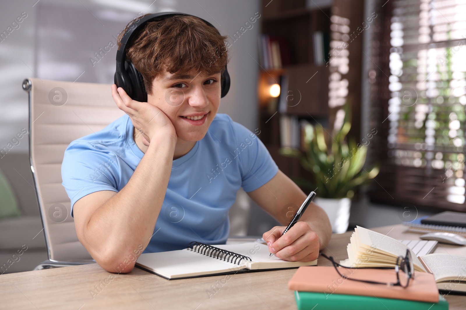 Teenage student in headphones taking notes while studying at table indoors Photo of Teenage student in headphones taking notes while studying at table indoors