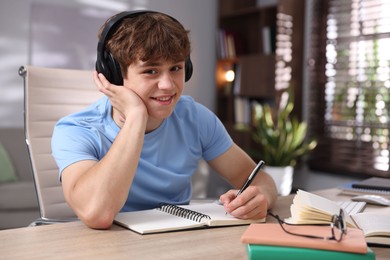 Photo of Teenage student in headphones taking notes while studying at table indoors