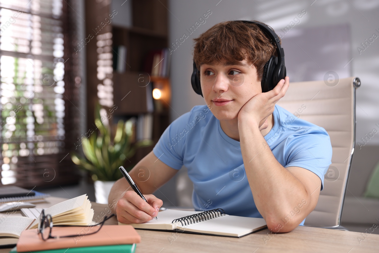 Teenage student in headphones taking notes while studying at table indoors Photo of Teenage student in headphones taking notes while studying at table indoors