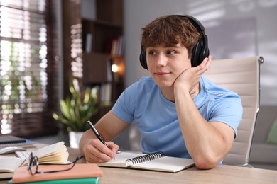 Photo of Teenage student in headphones taking notes while studying at table indoors