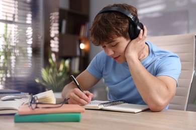 Photo of Teenage student in headphones taking notes while studying at table indoors
