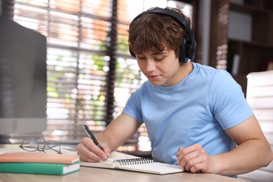 Photo of Teenage student in headphones taking notes while studying at table indoors