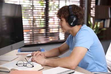 Photo of Teenage student with headphones using computer while studying at table indoors