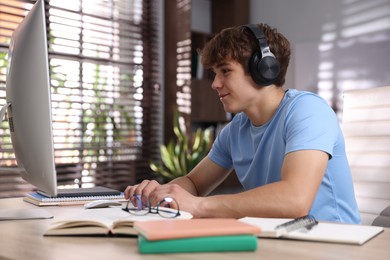 Photo of Teenage student with headphones using computer while studying at table indoors