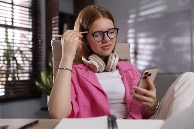 Photo of Teenage student with headphones using smartphone while studying at home