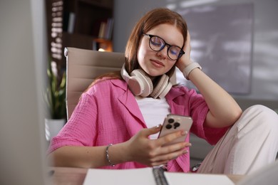 Photo of Teenage student with headphones using smartphone while studying at home