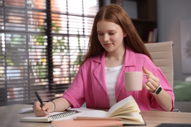 Photo of Teenage student with cup of drink taking notes while studying at table indoors