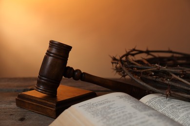 Photo of Judge's gavel, crown of thorns and Bible on wooden table against pale orange gradient background, closeup
