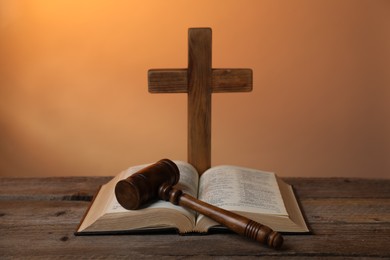 Photo of Judge's gavel, Bible and cross on wooden table against pale orange background, closeup