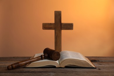 Photo of Judge's gavel, Bible and cross on wooden table against pale orange background, closeup