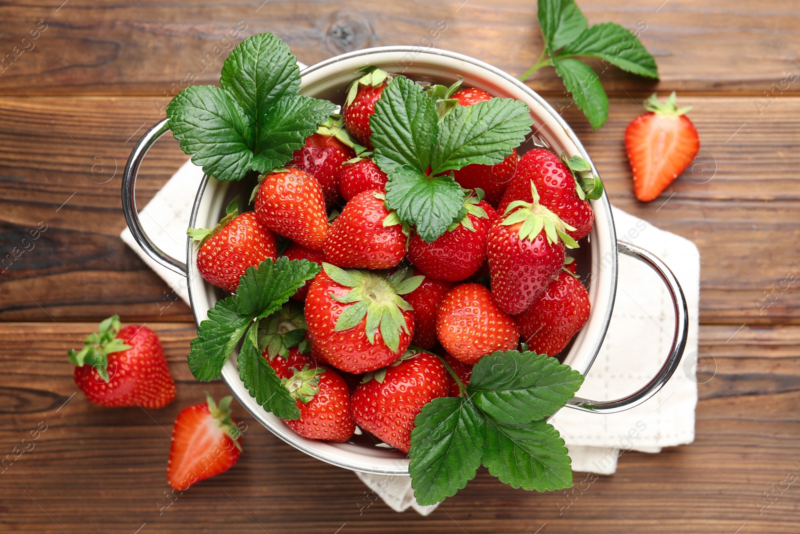 Fresh ripe strawberries and leaves in metal colander on wooden table, flat lay Photo of Fresh ripe strawberries and leaves in metal colander on wooden table, flat lay