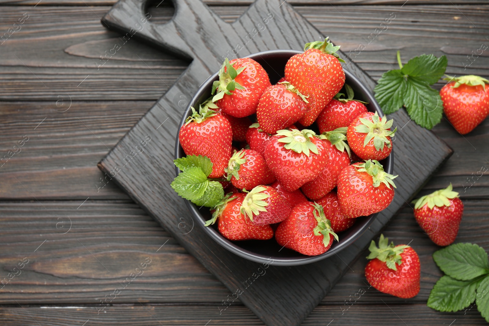 Fresh ripe strawberries and leaves in bowl on black wooden table, flat lay Photo of Fresh ripe strawberries and leaves in bowl on black wooden table, flat lay