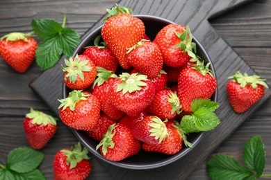 Photo of Fresh ripe strawberries and leaves in bowl on black wooden table, flat lay