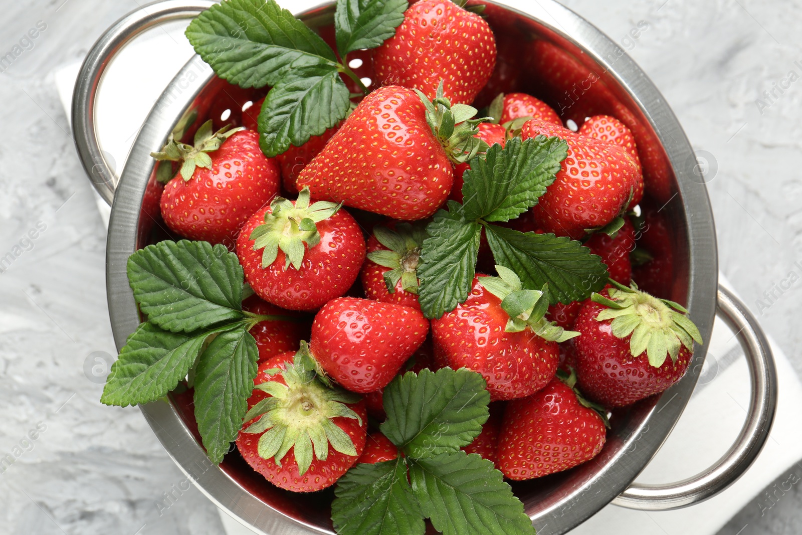 Delicious ripe strawberries and green leaves in colander on grey table, top view Photo of Delicious ripe strawberries and green leaves in colander on grey table, top view