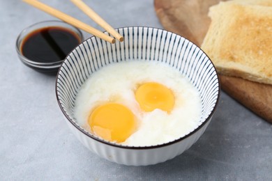 Photo of Traditional asian breakfast with half-boiled eggs in bowl, soy sauce, bread and chopsticks on grey table, above view