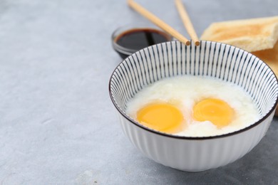 Photo of Traditional asian breakfast with half-boiled eggs in bowl, soy sauce, bread and chopsticks on grey table, closeup. Space for text