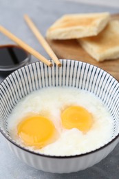 Photo of Traditional asian breakfast with half-boiled eggs in bowl, soy sauce, bread and chopsticks on grey table, closeup