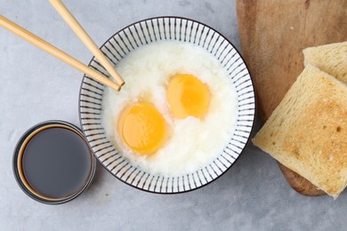 Photo of Traditional asian breakfast with half-boiled eggs in bowl, soy sauce, bread and chopsticks on grey table, flat lay