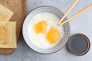 Photo of Traditional asian breakfast with half-boiled eggs in bowl, soy sauce, bread and chopsticks on grey table, flat lay
