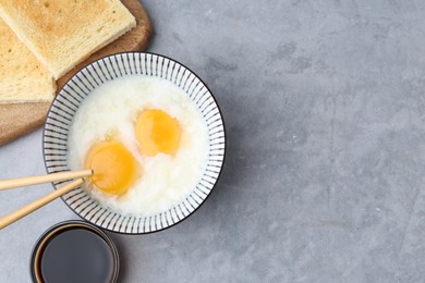 Photo of Traditional asian breakfast with half-boiled eggs in bowl, soy sauce, bread and chopsticks on grey table, flat lay. Space for text